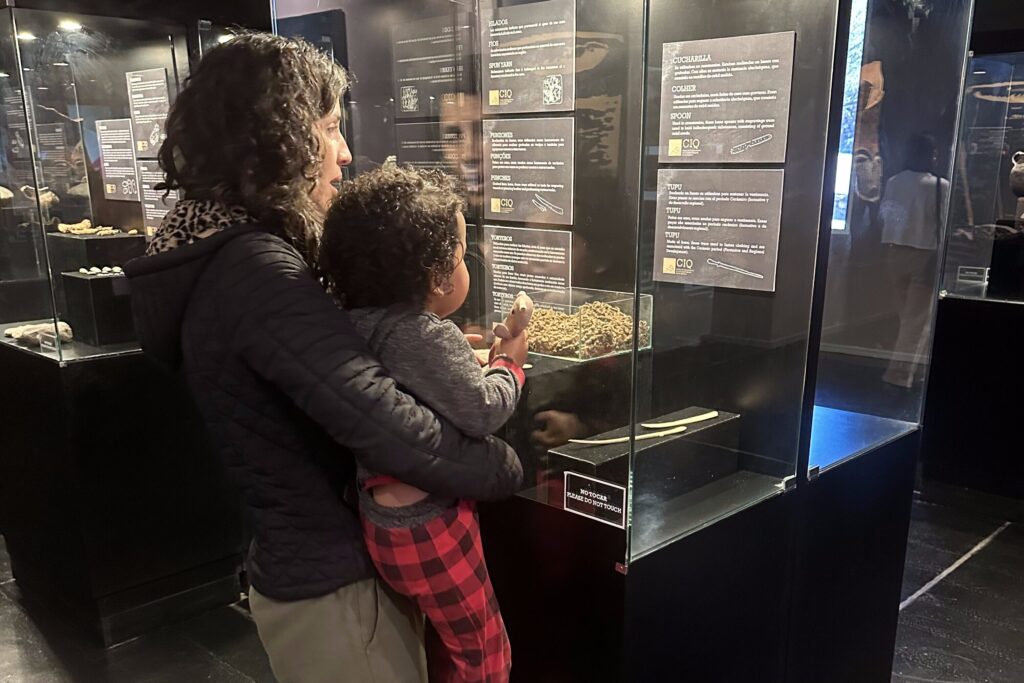 mulher com uma crinaça no colo observando um expositor com objetos arqueológicos num museu