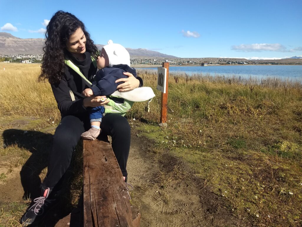 mulher centada num banco de madeira com uma criança de colo. Os dois de olham apaixonados. Todos estão agasalhados, pois apesar do sol está bem frio.
