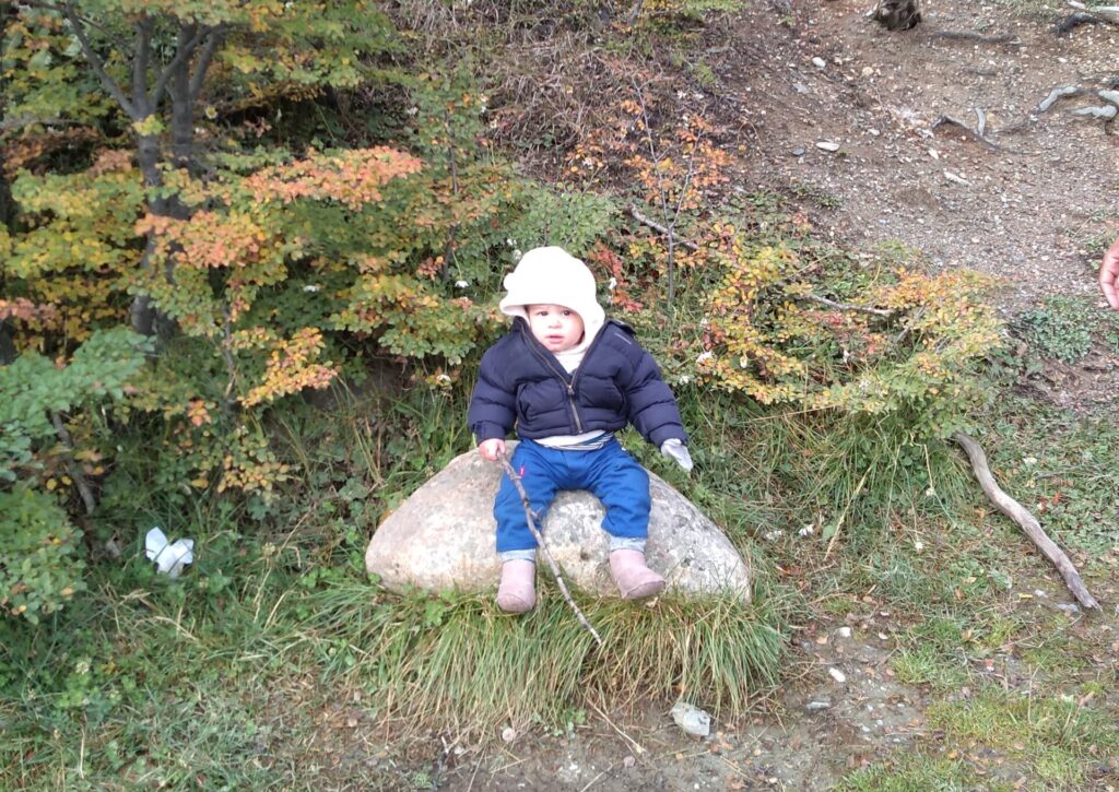 crianaça de um ano sentado numa pedra com um graveto na mão, parece estar frio pois ela está bem agasalhada.