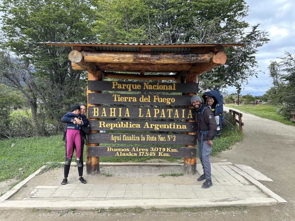 foto de uma família: pai, mãe e um casal de filhos pequeno na frente da placa da da baia de lapataia e fimnal da ruta 3 no parque do fim do mundo